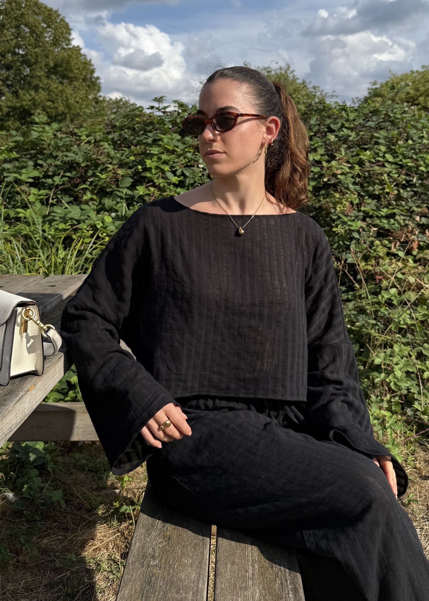 A woman wearing a black striped long-sleeve top and matching wide-leg trousers sits on a wooden bench outdoors. A white handbag with gold hardware rests on the bench beside her.