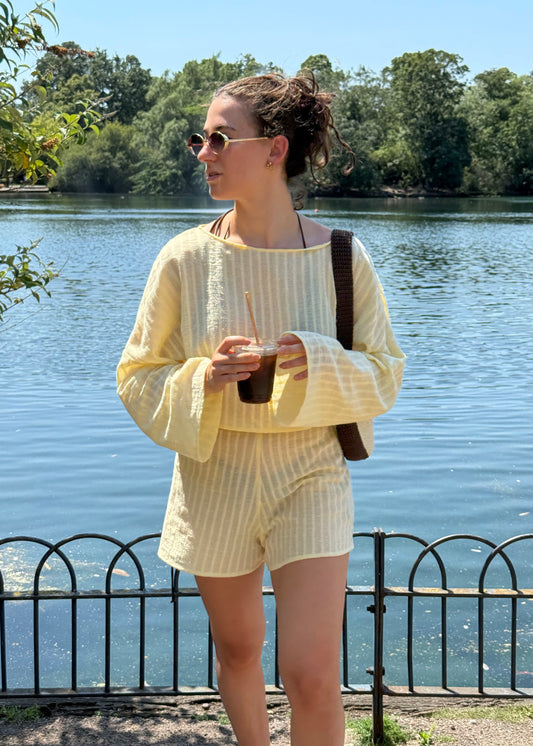 A woman wearing a pale yellow two-piece set with a long-sleeve top and matching shorts, standing outdoors by a lake with trees in the background.