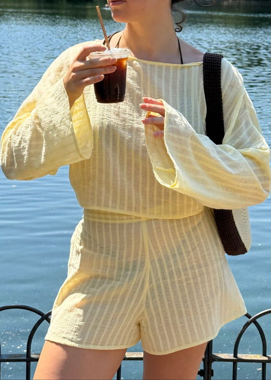 A woman wearing a pale yellow and white vertically striped two-piece set with a boat neckline, long flared sleeves, and matching shorts, standing by a body of water and holding an iced coffee.