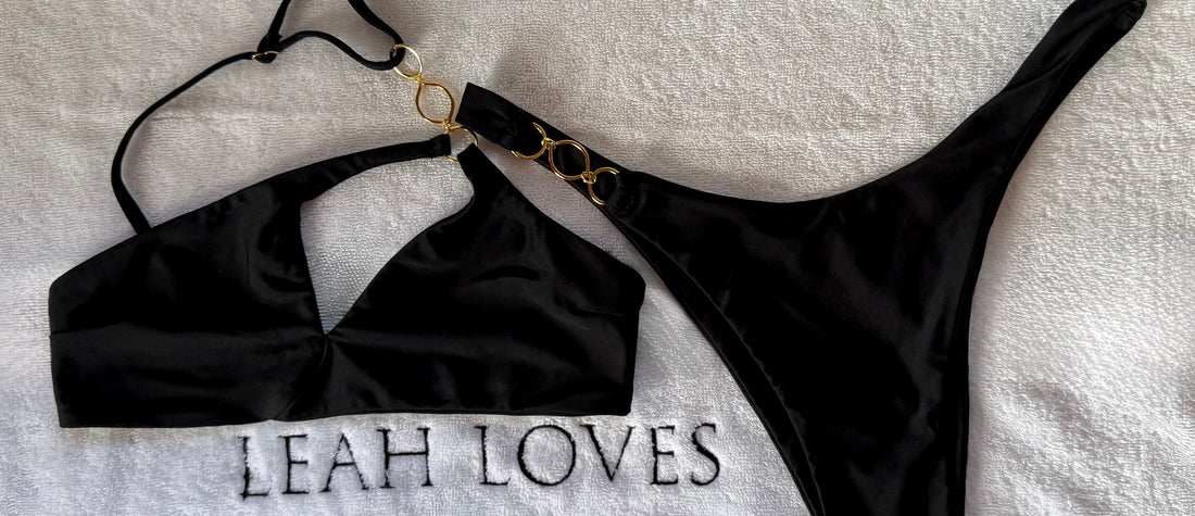 Leah Loves Ivy Asymmetric Bikini in Black with gold hardware photographed as a flat lay on a branded Leah Loves towel. 