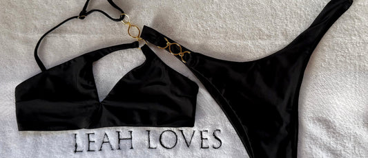 Leah Loves Ivy Asymmetric Bikini in Black with gold hardware photographed as a flat lay on a branded Leah Loves towel. 