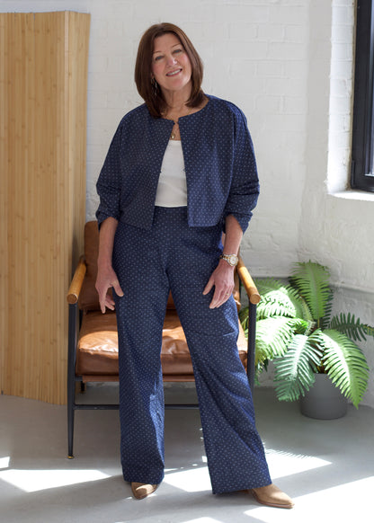 Woman in a blue patterned outfit standing in a room with a chair and plants.