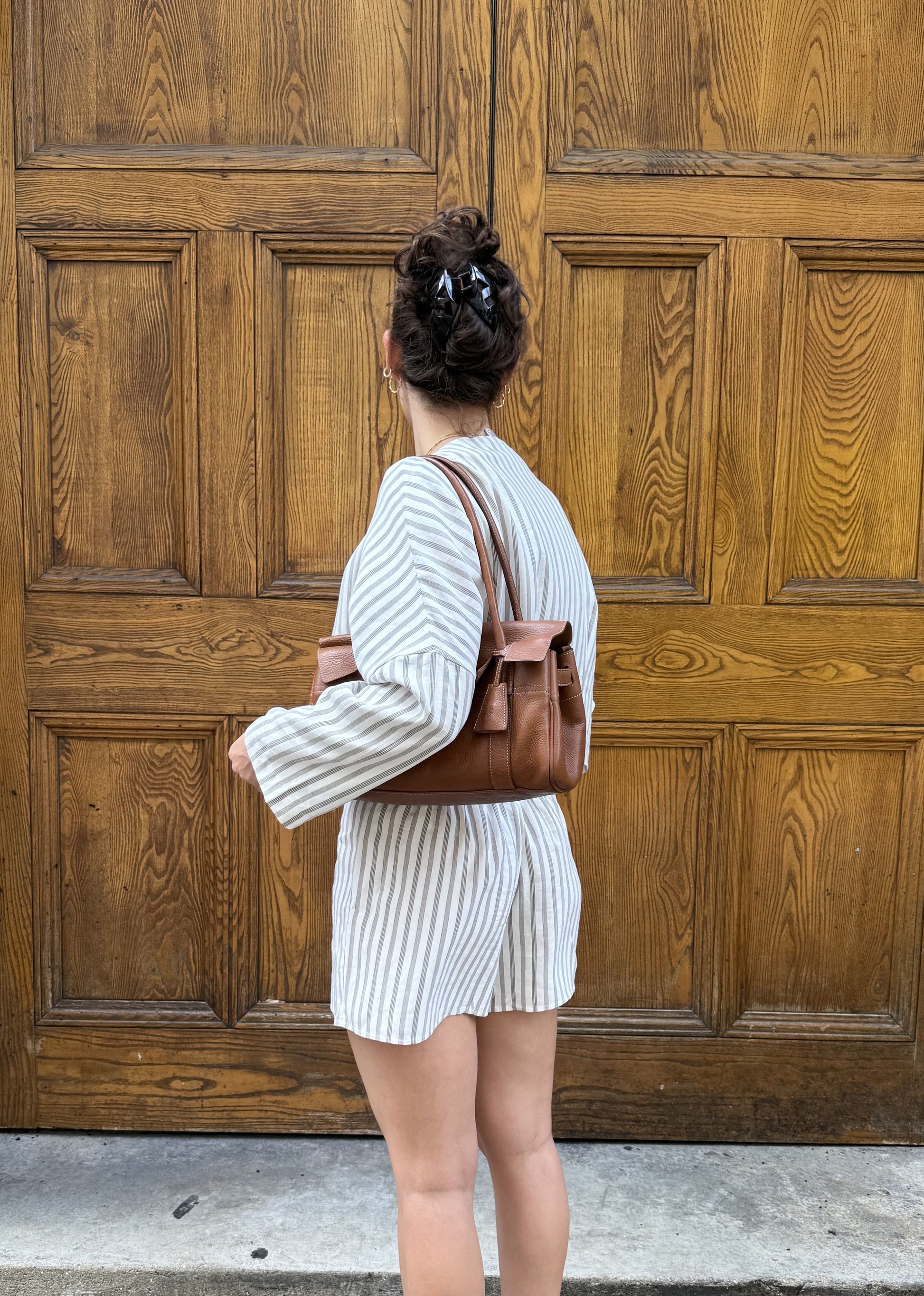 A woman stands with her back to the camera in front of large wooden double doors, wearing a white and brown vertically striped matching set and a brown leather shoulder bag.