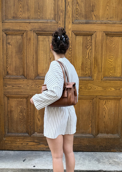 A woman stands with her back to the camera in front of large wooden double doors, wearing a white and brown vertically striped matching set and a brown leather shoulder bag.