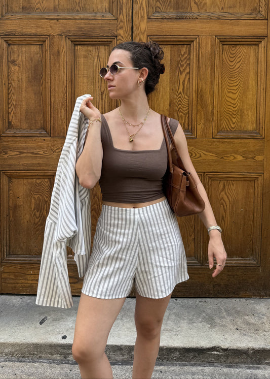 A woman stands in front of large wooden doors wearing a brown vest, white shorts with brown pinstripes, and holding a matching striped jacket. She has a brown shoulder bag and sunglasses.