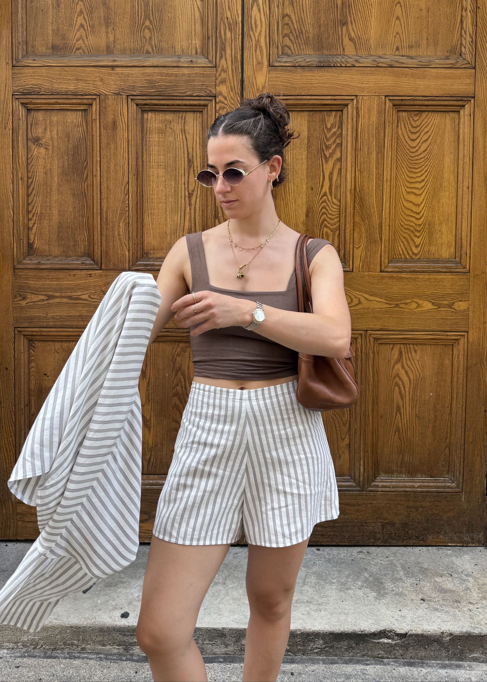 A woman stands in front of large wooden doors wearing a brown vest, white and brown vertically striped shorts, and holding a matching striped jacket. She has a brown shoulder bag and sunglasses.