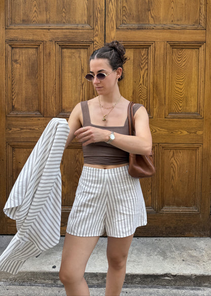 A woman stands in front of large wooden doors wearing a brown vest, white and brown vertically striped shorts, and holding a matching striped jacket. She has a brown shoulder bag and sunglasses.
