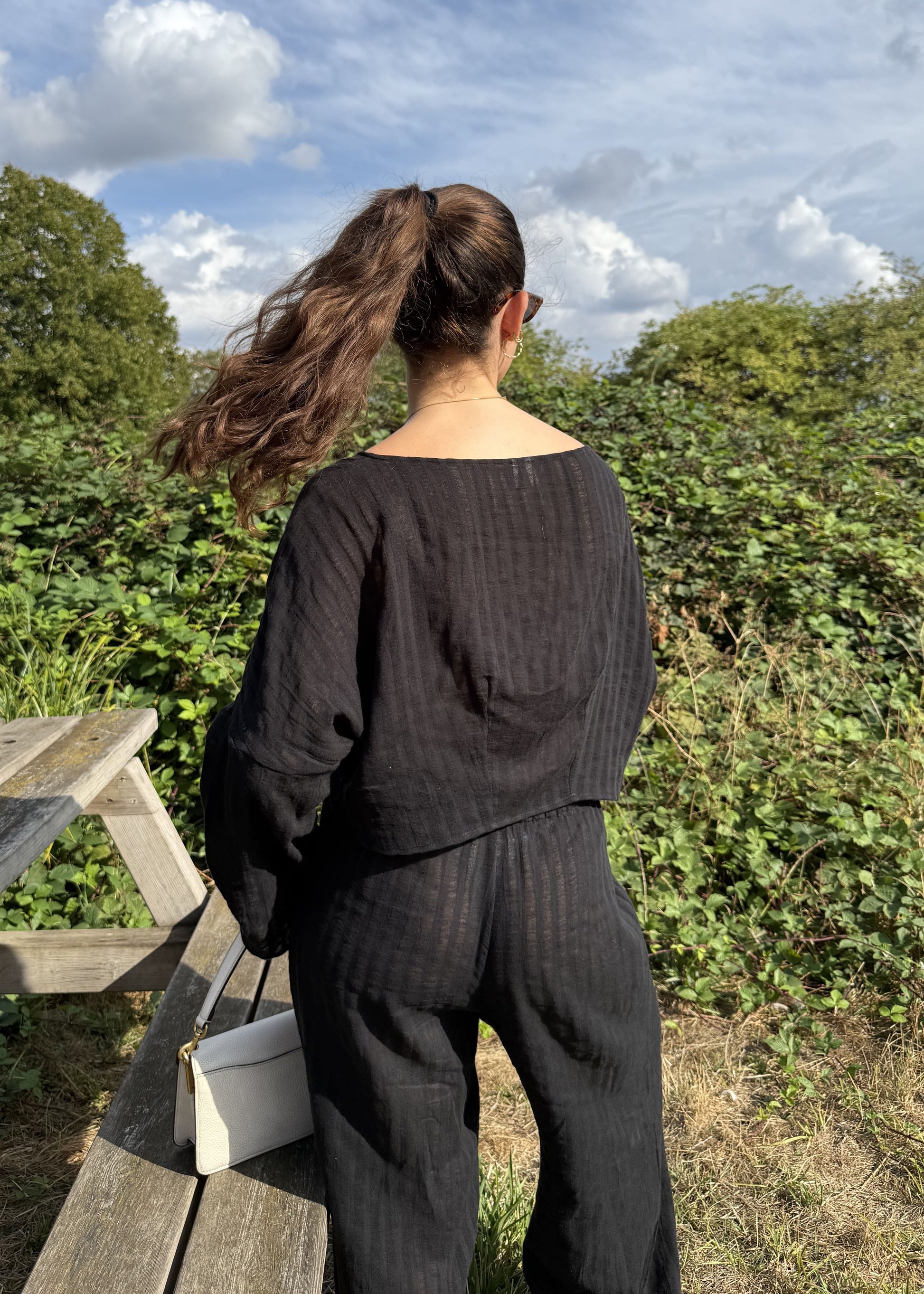 A woman seen from behind wearing a matching black two-piece outfit, standing outdoors next to a wooden picnic table with green bushes and a blue sky in the background.