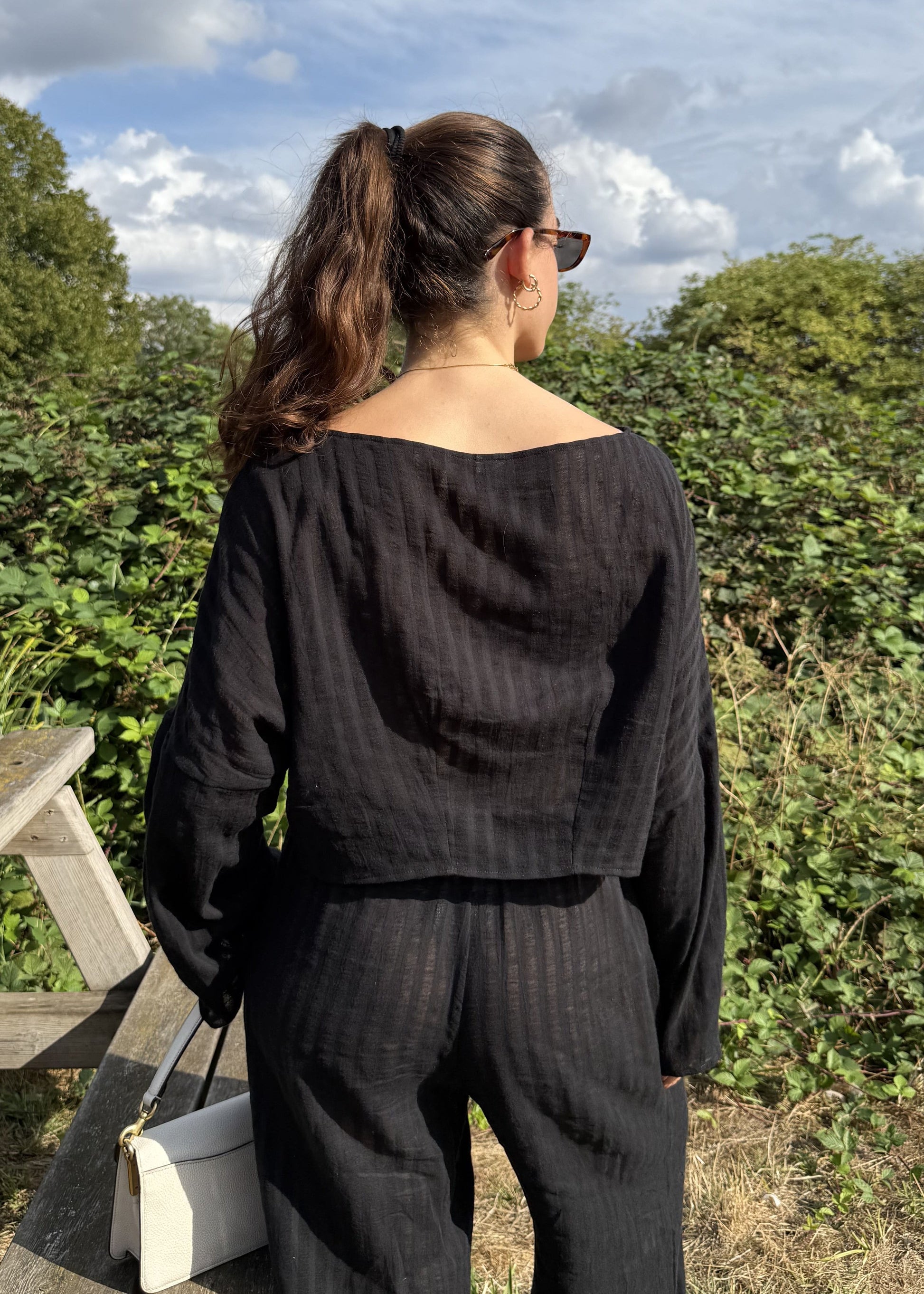 A woman viewed from behind wearing a matching black long-sleeve top and wide-leg trousers, standing outdoors on a wooden bench with green bushes and a blue sky in the background.