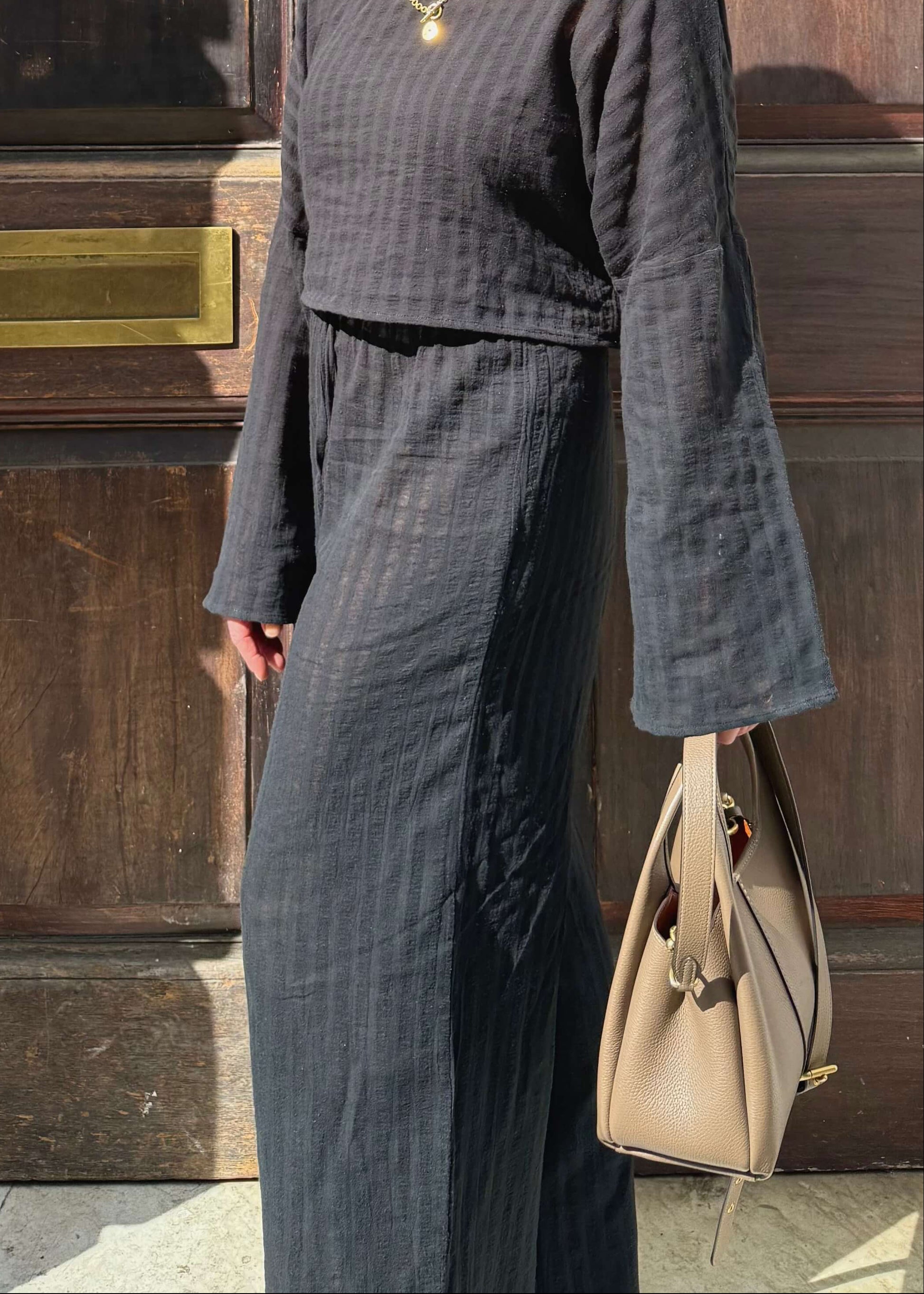 A woman wearing a black striped two-piece set with long bell sleeves and wide-leg pants, standing in front of a large dark wooden door and holding a handbag.