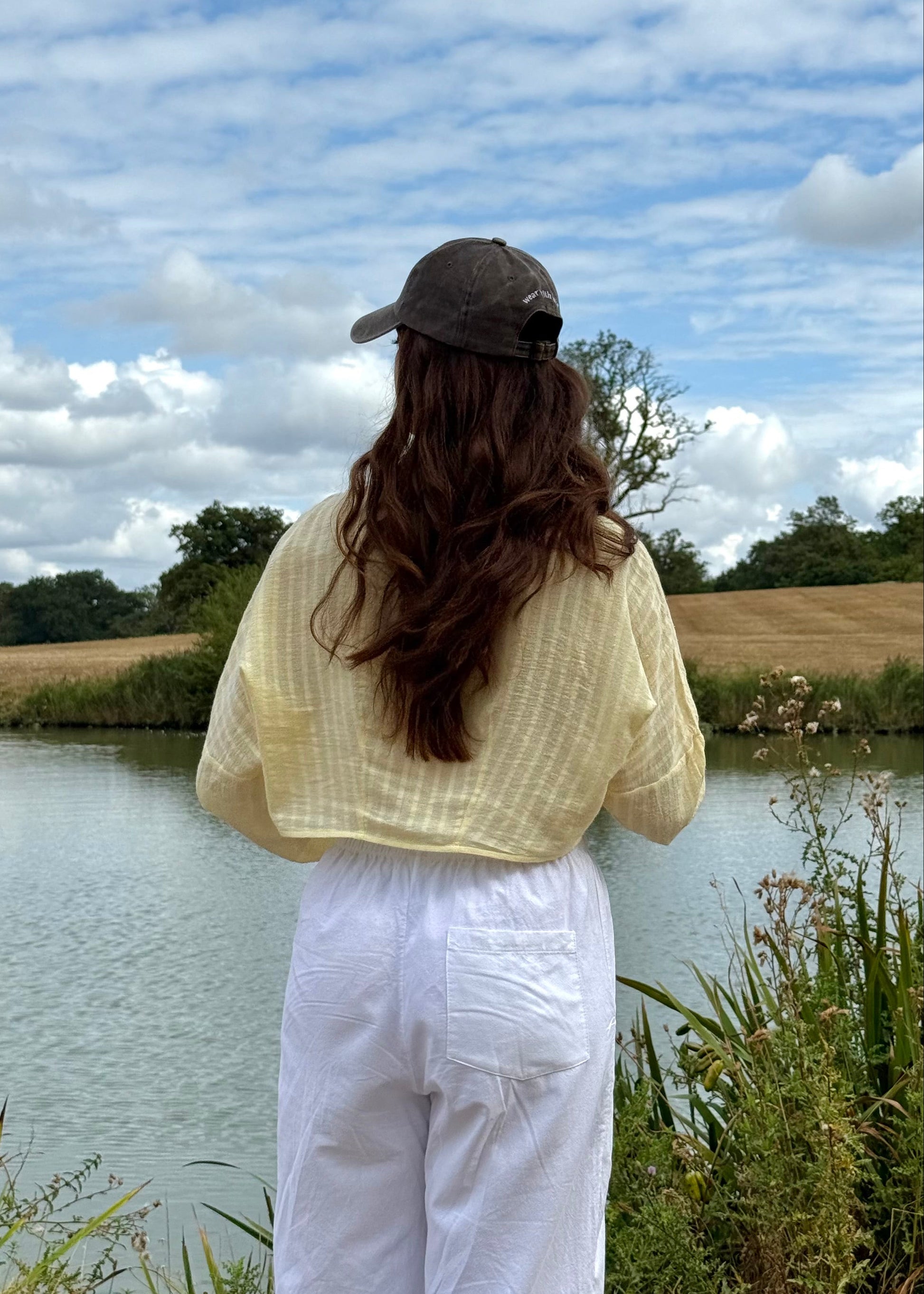 A woman stands with her back to the camera by a calm lake, wearing a pale yellow top, white pants, and a brown cap, with a grassy field and cloudy blue sky in the background.