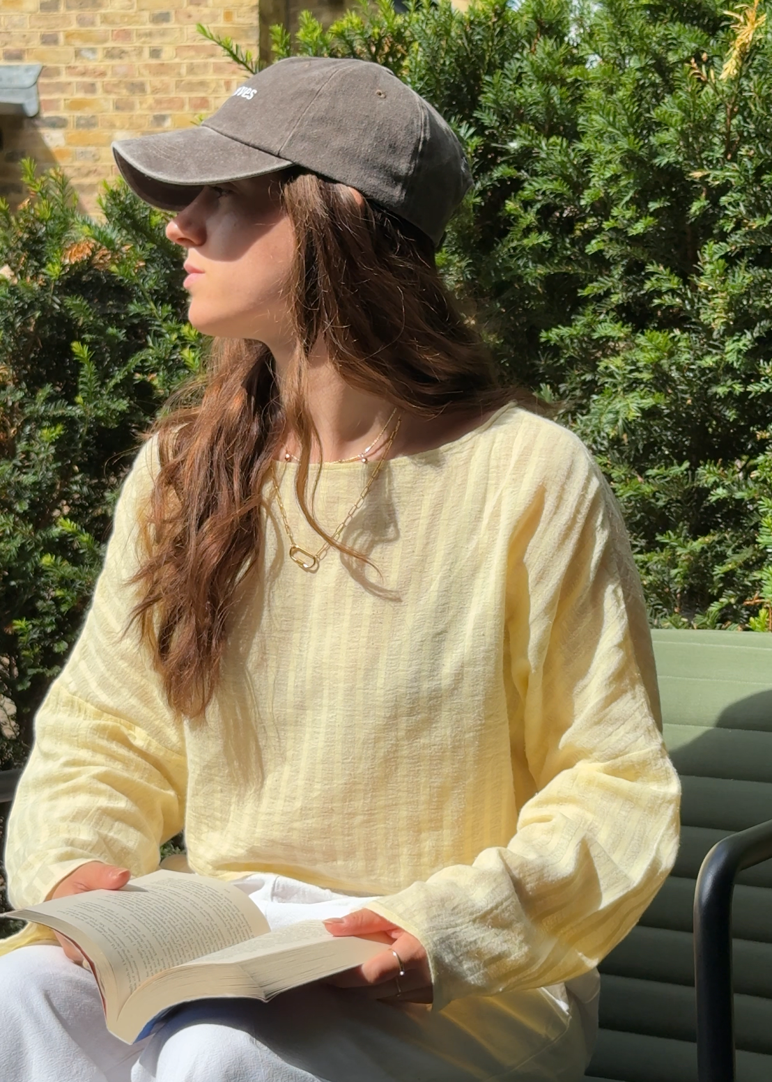 A woman is wearing a pale yellow long-sleeve top whilst sitting in the sun with a book on her lap. 