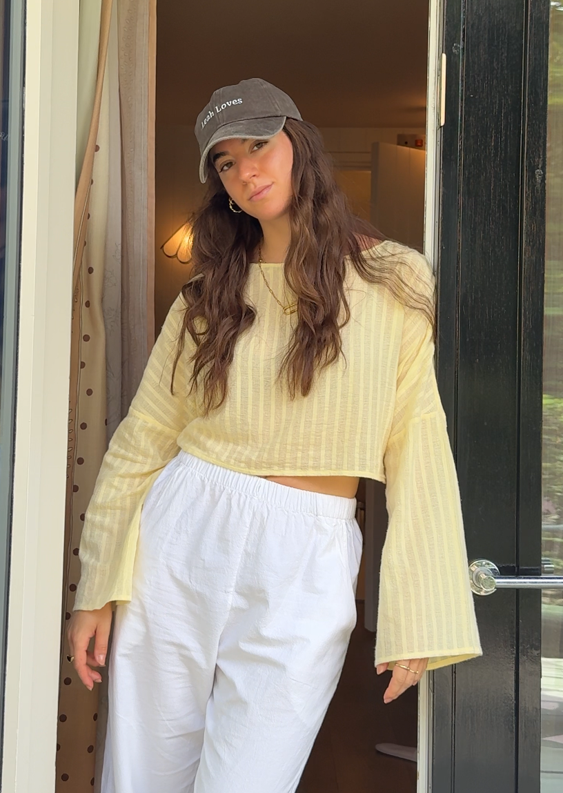 A woman stands in an open doorway wearing a pale yellow striped long-sleeve top with flared bell sleeves, white wide-leg pants, and a brown Leah Loves cap.