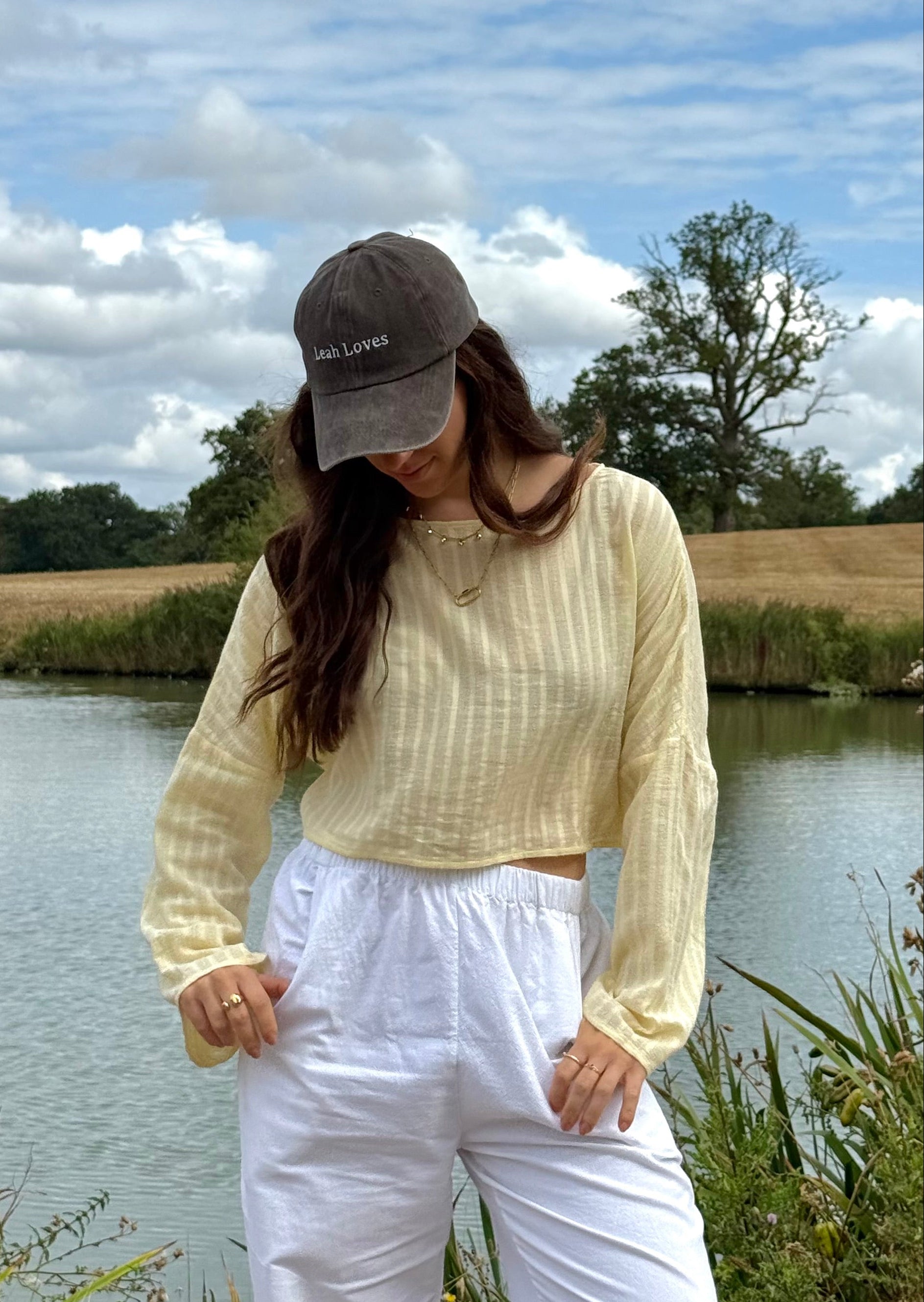 A woman stands outdoors by a river wearing a pale yellow striped long-sleeve top, white pants, and a brown cap with white text that reads 'Leah Loves'.
