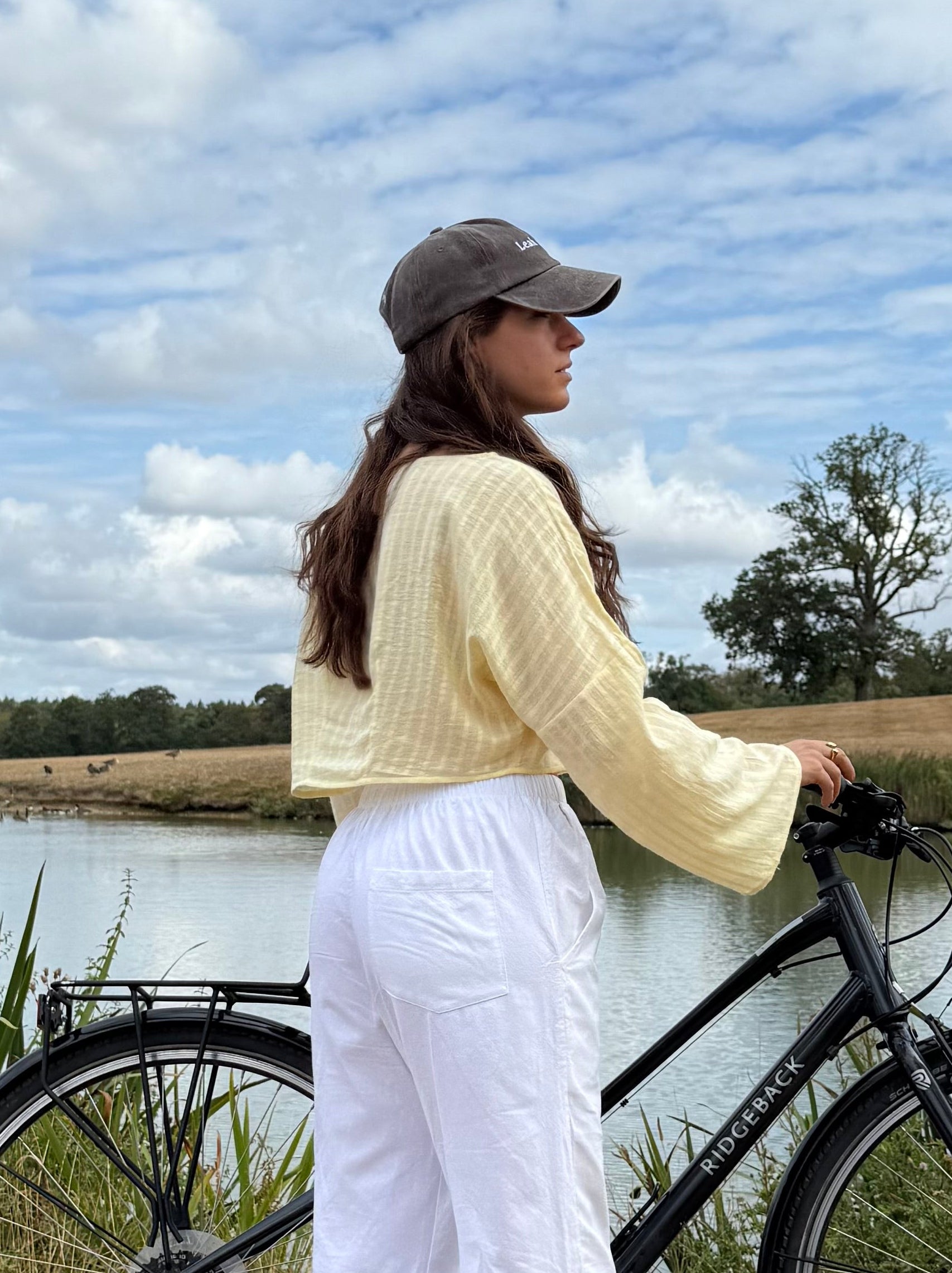 A woman stands outdoors beside a black bicycle near a lake, wearing a pale yellow long sleeve top, white wide-leg pants, and a brown cap. The background shows grass, trees, open land, and a blue sky with clouds.