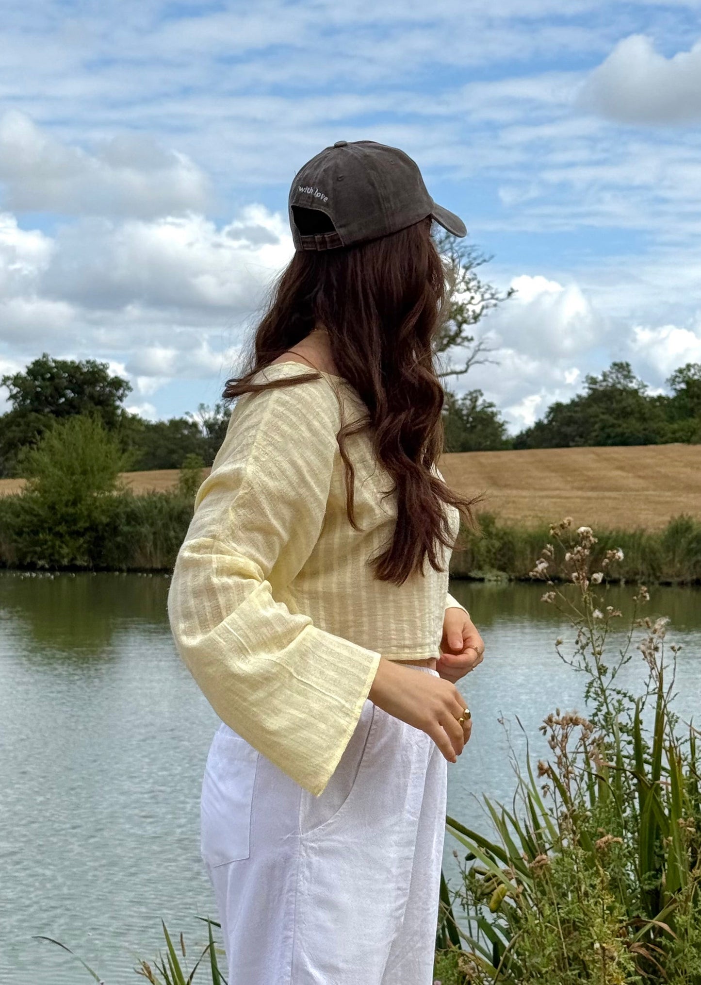 A woman stands outdoors by a lake wearing a pale yellow long-sleeve top, white wide-leg pants, and a brown cap, with grass, water, and a cloudy sky in the background.