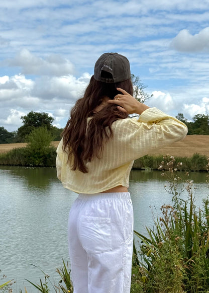 A woman stands with her back to the camera by a lake, wearing a pale yellow striped long-sleeve top, white trousers, and a dark down cap against a backdrop of water, grass, and a cloudy blue sky