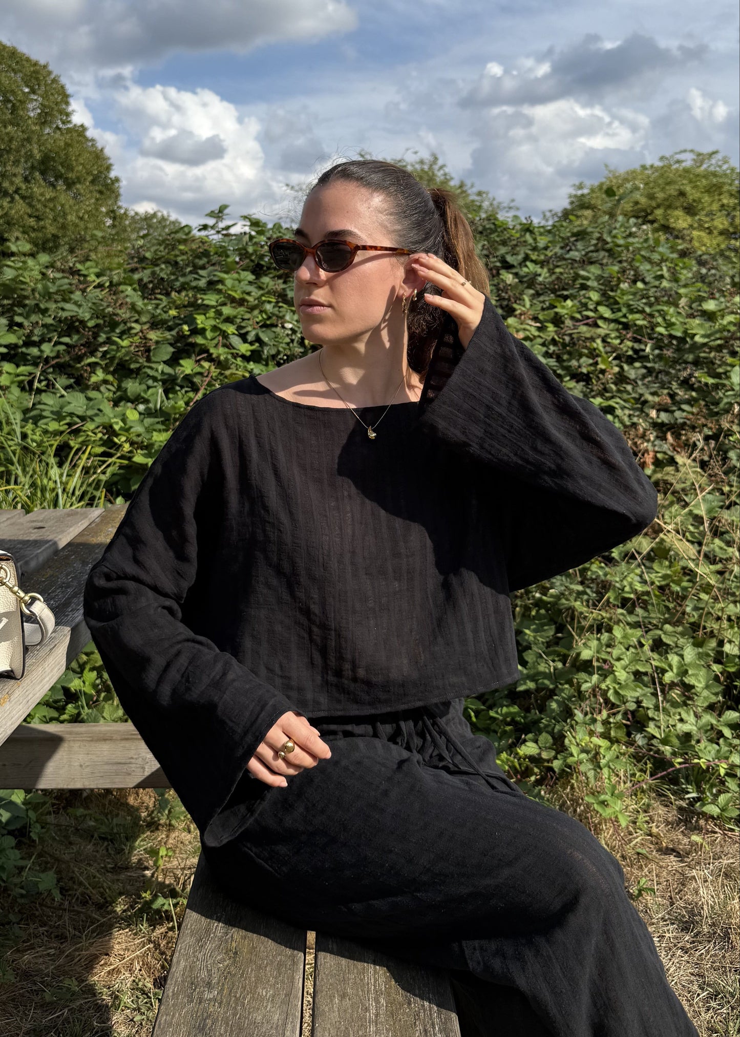 A woman wearing a black long-sleeve top and black trousers sits on a wooden bench outdoors, with a cream handbag resting on the bench beside her. The background features green foliage and a blue sky with clouds.