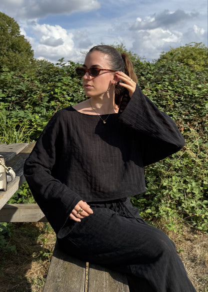A woman wearing a black long-sleeve top and black trousers sits on a wooden bench outdoors, with a cream handbag resting on the bench beside her. The background features green foliage and a blue sky with clouds.