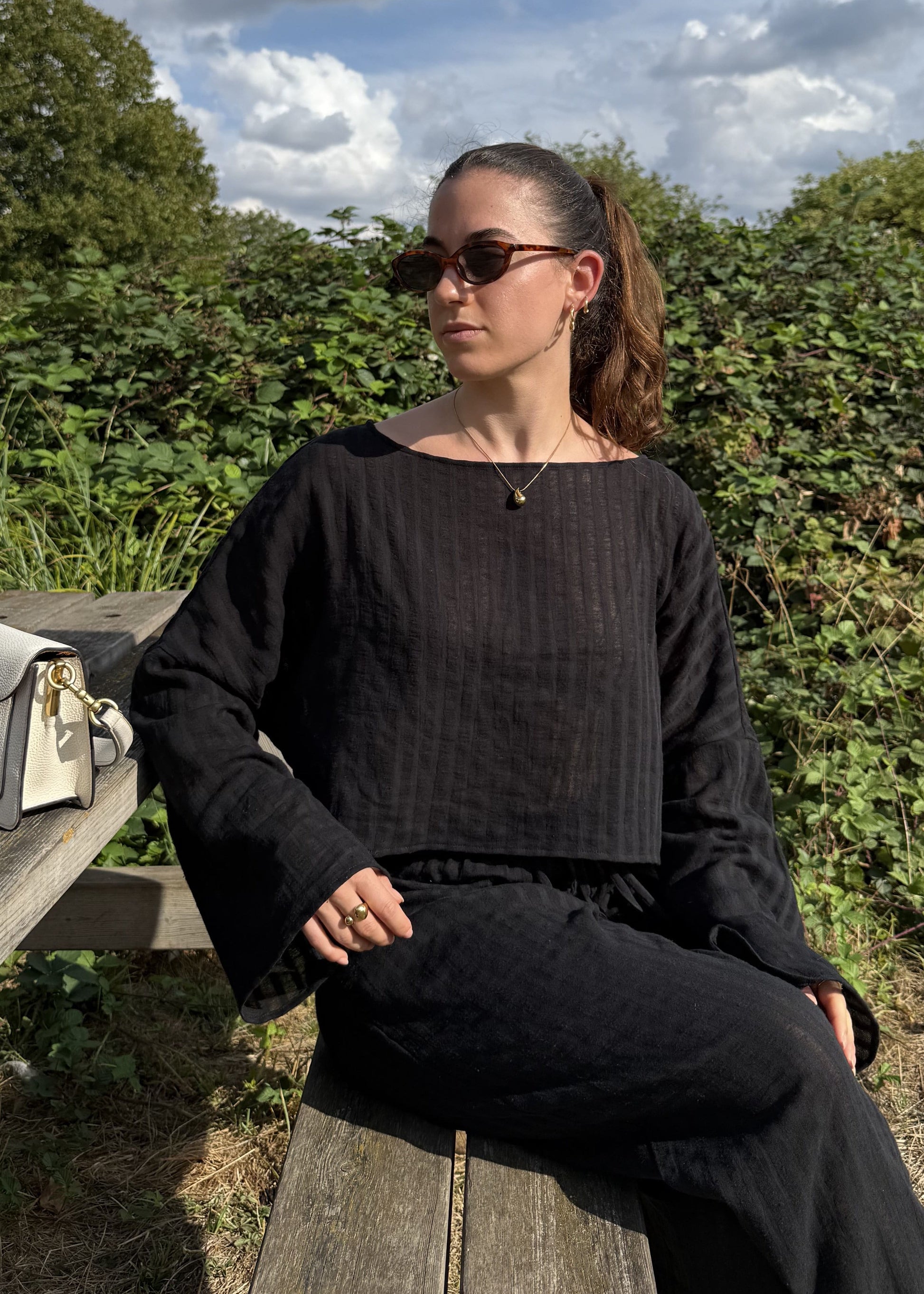 A woman wearing a black striped long-sleeve top and matching wide-leg trousers sits on a wooden bench outdoors. A white handbag with gold hardware rests on the bench beside her.