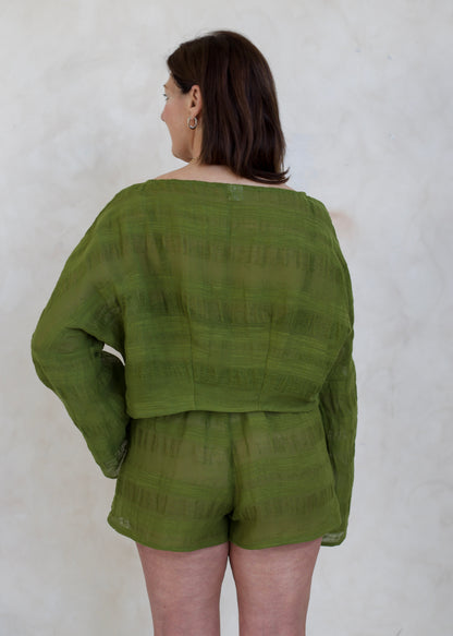 A woman shown from the back wearing a matching olive green long-sleeve top and shorts set against a textured off-white wall.