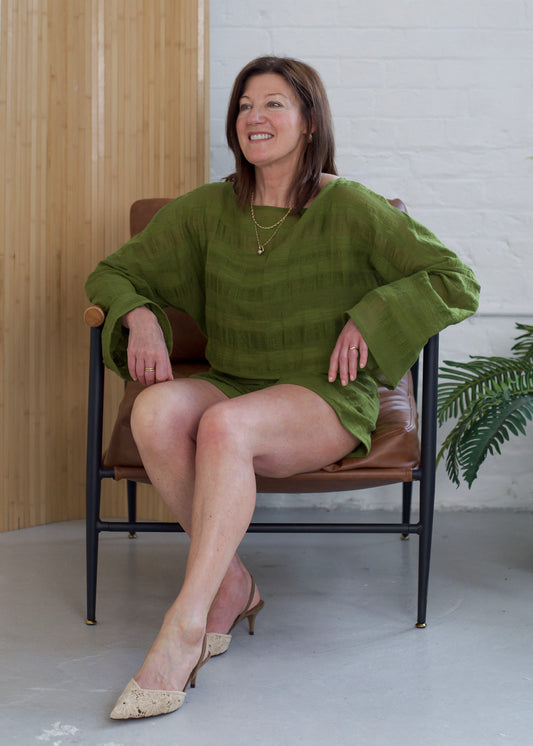 A woman wearing a textured olive green long-sleeve top and matching shorts sits in a brown leather armchair with a black metal frame, set against a background of a light wood panel, white brick wall, and a potted plant.