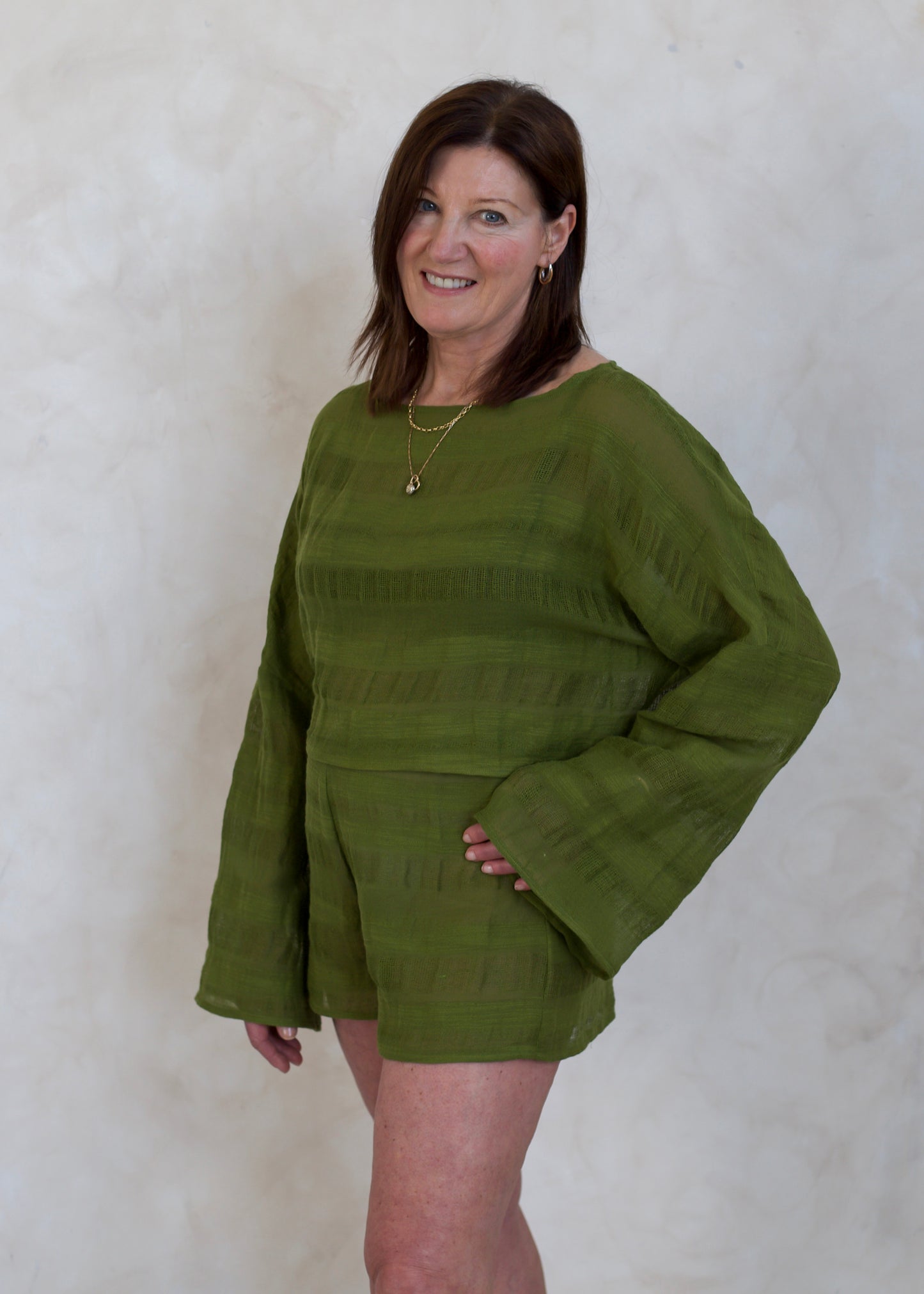 A woman modeling a matching olive green long-sleeve top and shorts set against a textured off-white wall.