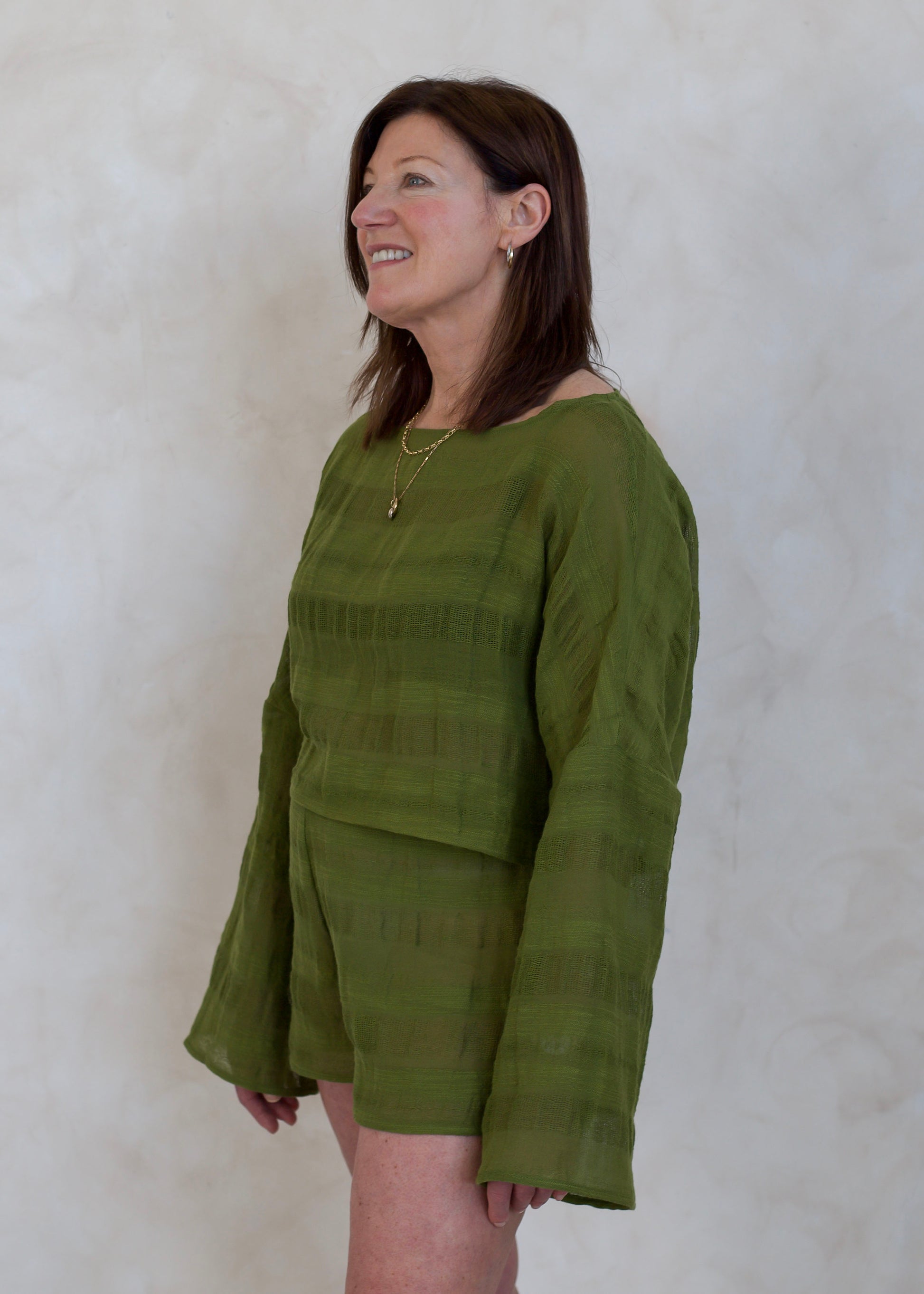 A woman wearing a textured olive green two-piece set consisting of a long-sleeve top and matching shorts, standing against a neutral textured wall.
