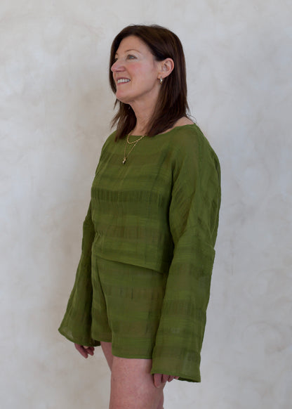 A woman wearing a textured olive green two-piece set consisting of a long-sleeve top and matching shorts, standing against a neutral textured wall.