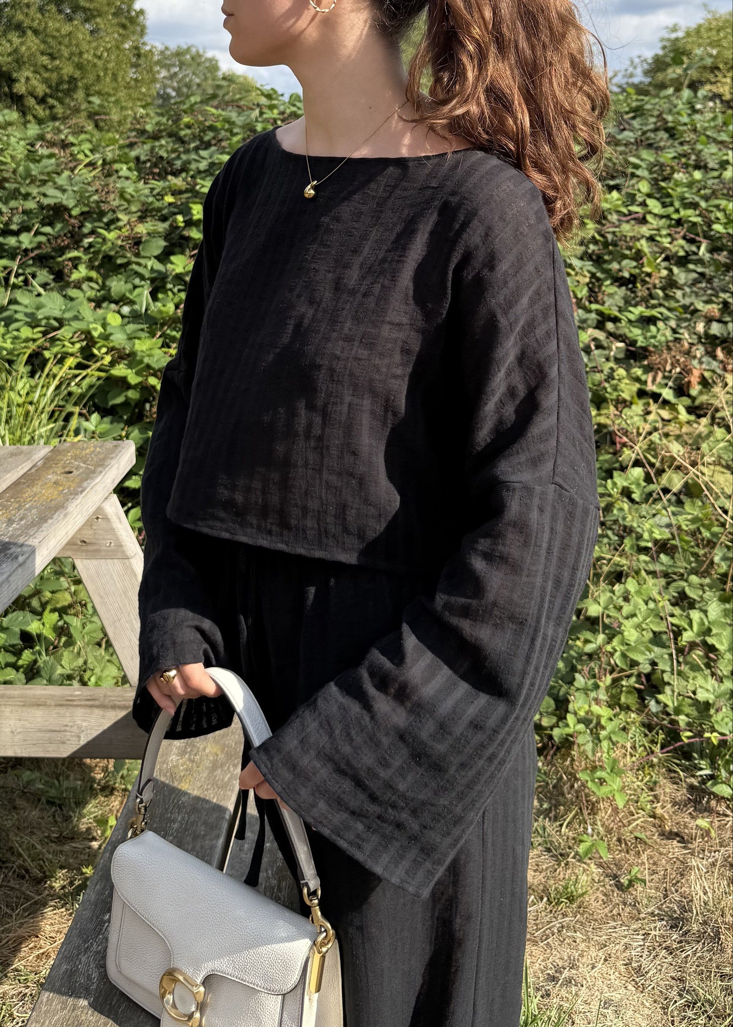 A woman with her hair in a ponytail stands outdoors next to a wooden picnic table, wearing a black striped long-sleeve top and matching trousers, holding a cream handbag. The background features green bushes and a blue sky with white clouds.