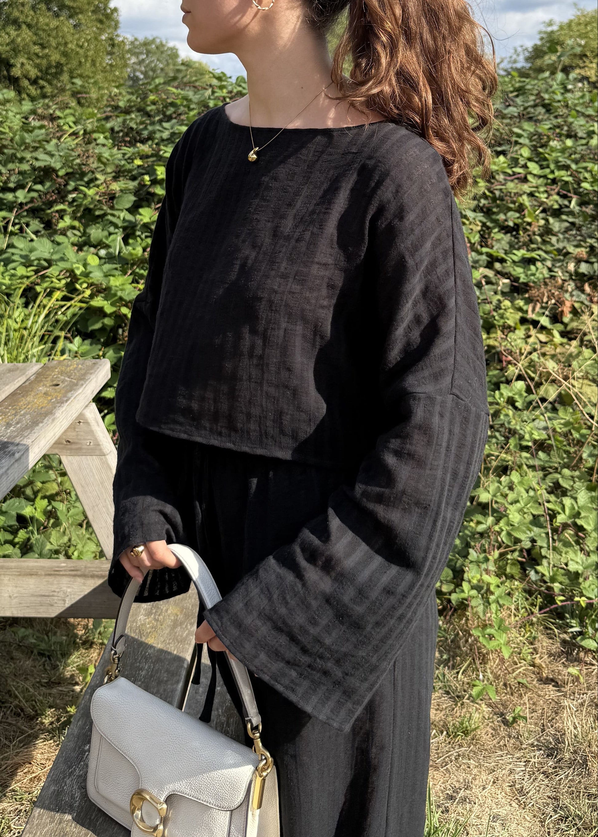 A woman with her hair in a ponytail stands outdoors next to a wooden picnic table, wearing a black striped long-sleeve top and matching trousers, holding a cream handbag. The background features green bushes and a blue sky with white clouds.