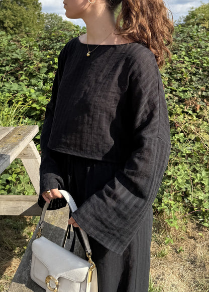 A woman with her hair in a ponytail stands outdoors next to a wooden picnic table, wearing a black striped long-sleeve top and matching trousers, holding a cream handbag. The background features green bushes and a blue sky with white clouds.