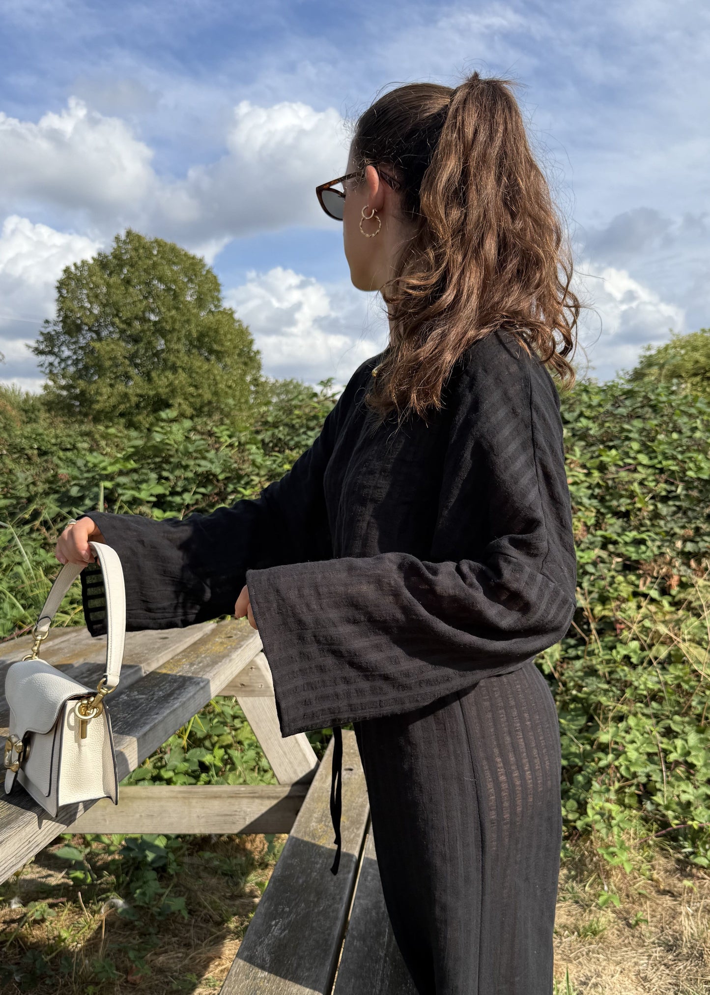 A woman wearing a black striped long-sleeve top and matching wide-leg trousers stands outdoors next to a wooden bench, holding a small cream-colored handbag. The background features green hedges and a blue sky with white clouds.