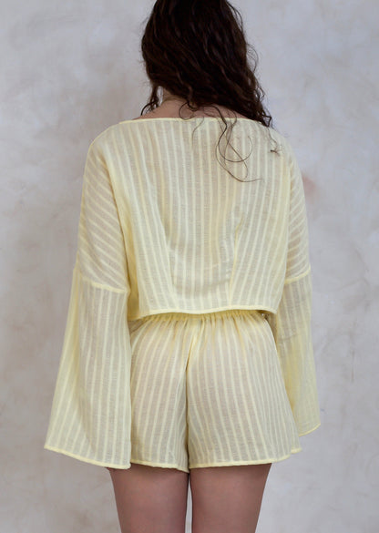 A model viewed from behind wearing a pale yellow vertically striped two-piece set consisting of a long-sleeve top and matching shorts, set against a textured light gray wall.