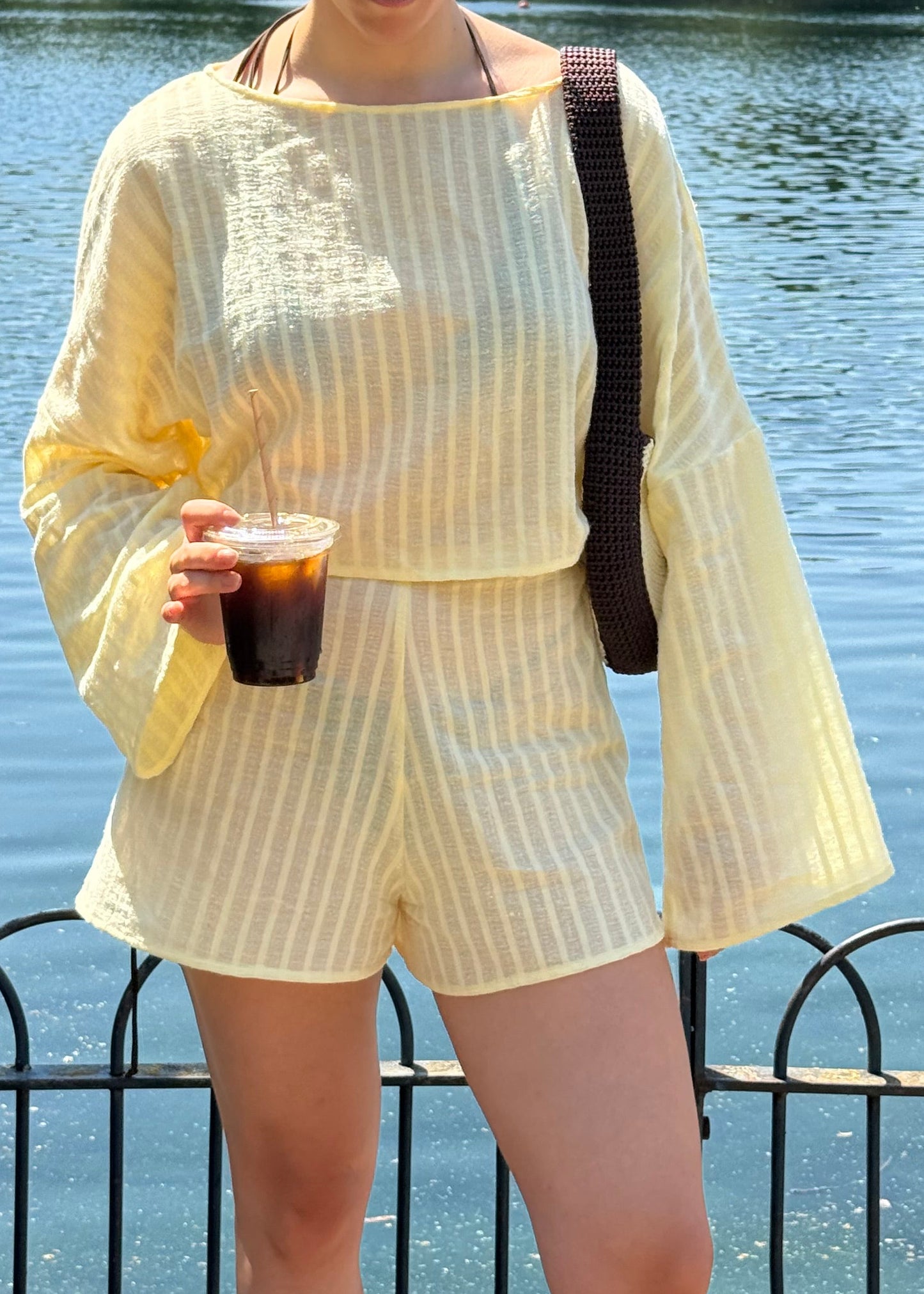 A person wearing a pale yellow two-piece set with vertical stripes, featuring a long-sleeve top and matching shorts, standing outdoors by a body of water and holding an iced coffee.