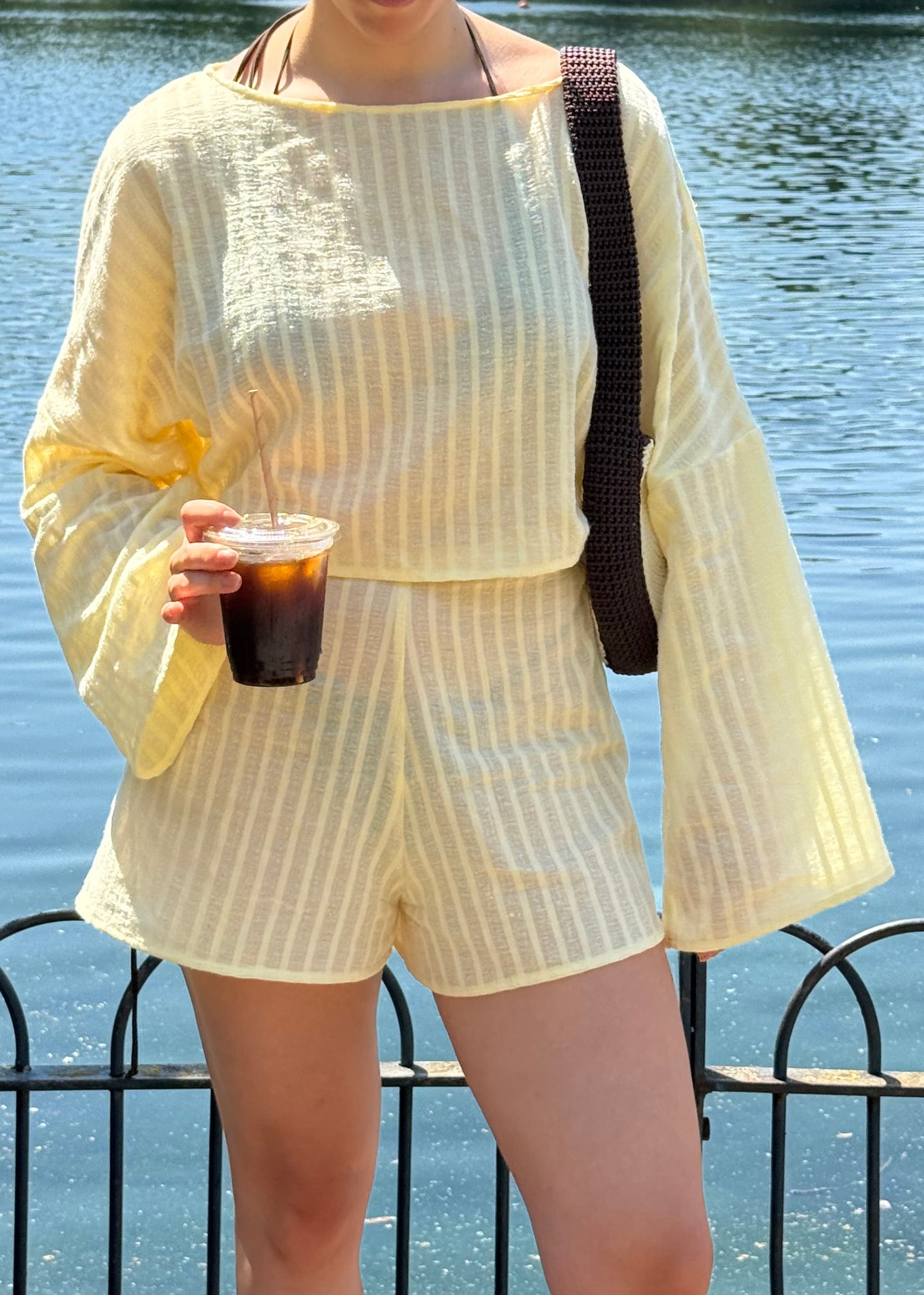 A person wearing a pale yellow two-piece set with vertical stripes, featuring a long-sleeve top and matching shorts, standing outdoors by a body of water and holding an iced coffee.
