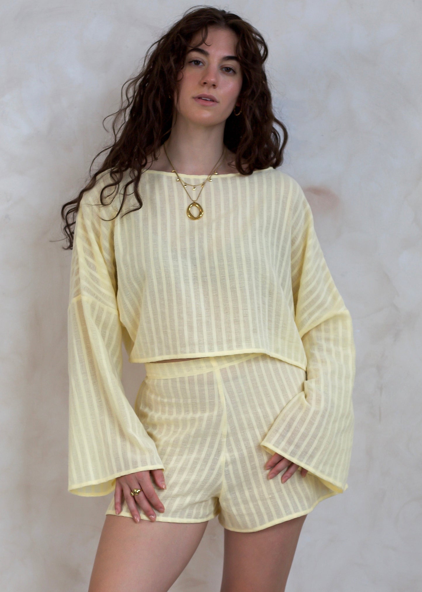 A woman modeling a pale yellow and white vertically striped two-piece set consisting of a long-sleeve cropped top and matching shorts, standing against a textured light gray wall.