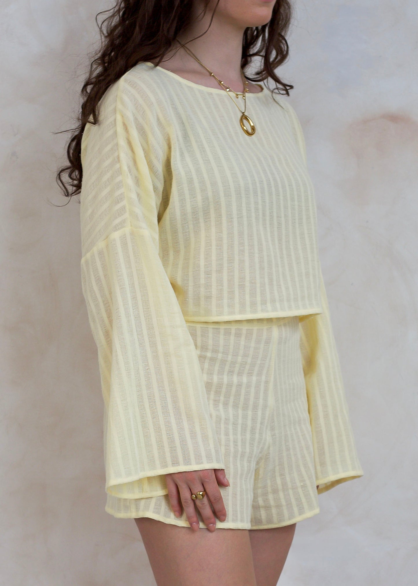 A woman wearing a pale yellow two-piece set with vertical textured stripes, featuring a long-sleeve top and matching shorts, set against a neutral textured wall.