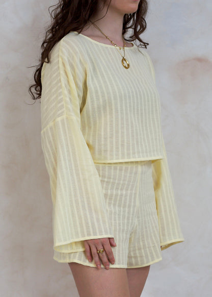 A woman wearing a pale yellow two-piece set with vertical textured stripes, featuring a long-sleeve top and matching shorts, set against a neutral textured wall.
