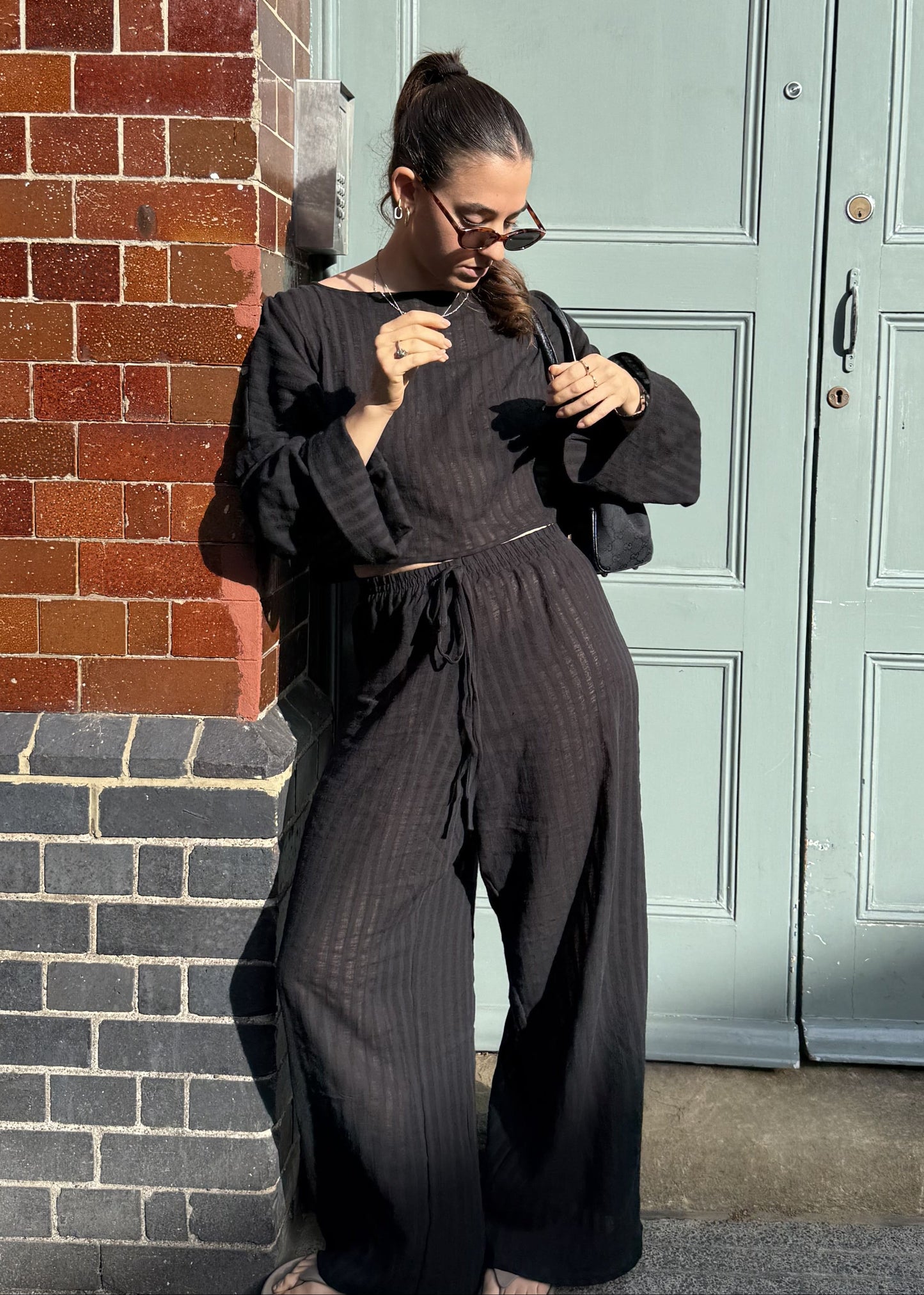 A woman wearing a black striped co-ord with wide leg trousers and long sleeves top stands on a sidewalk in front of a light green door and a red brick wall.