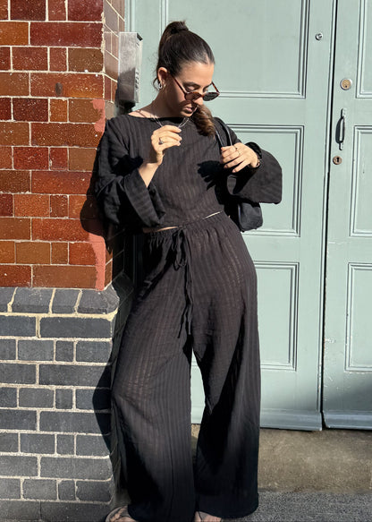 A woman wearing a black striped co-ord with wide leg trousers and long sleeves top stands on a sidewalk in front of a light green door and a red brick wall.