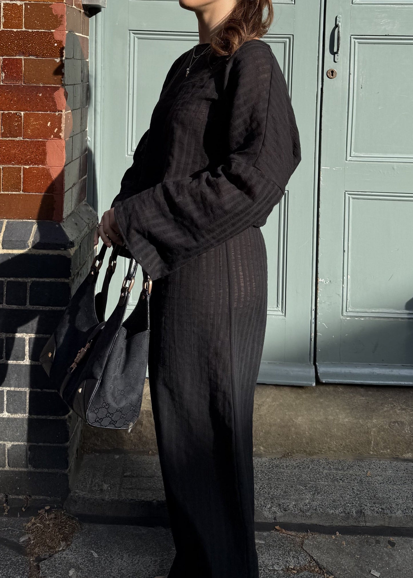 A woman wearing a black striped long-sleeve top and matching wide-leg trousers stands outdoors in front of a pale green door and red brick wall, holding a black handbag.