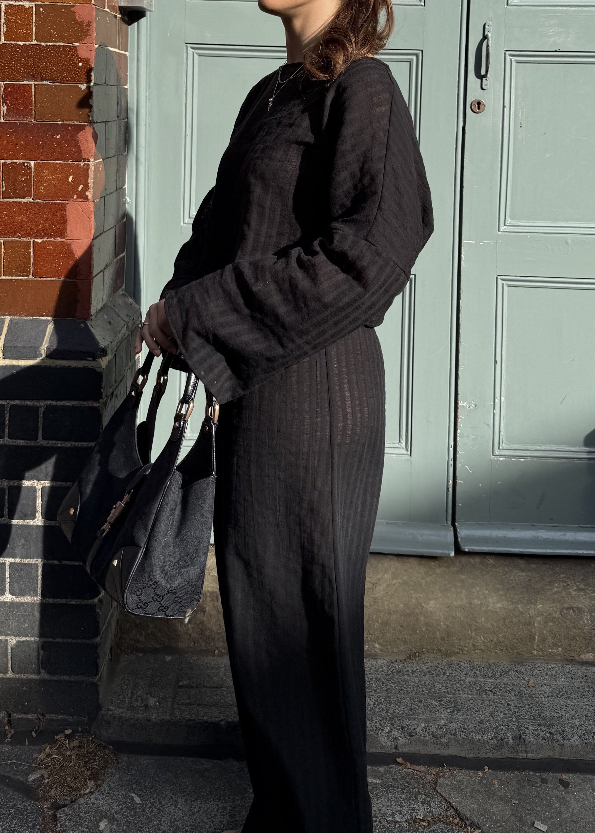 A woman wearing a black striped long-sleeve top and matching wide-leg trousers stands outdoors in front of a pale green door and red brick wall, holding a black handbag.