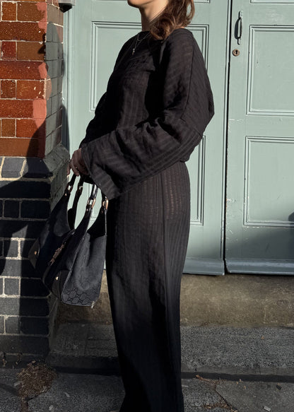 A woman wearing a black striped long-sleeve top and matching wide-leg trousers stands outdoors in front of a pale green door and red brick wall, holding a black handbag.