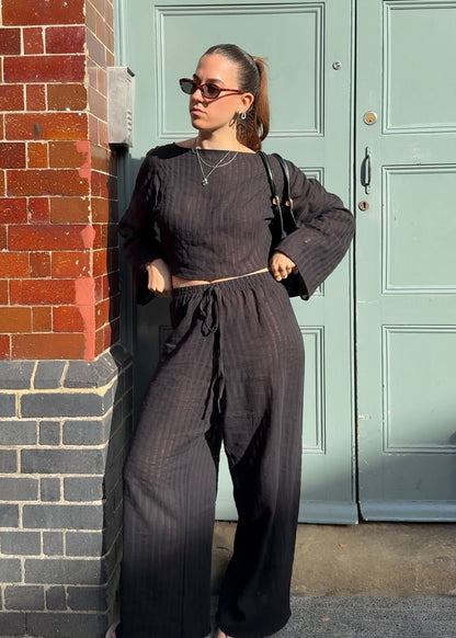 A woman wearing a black striped co-ord with a drawstring waist and wide-leg pants, standing outdoors next to a red brick pillar in front of a light green door.