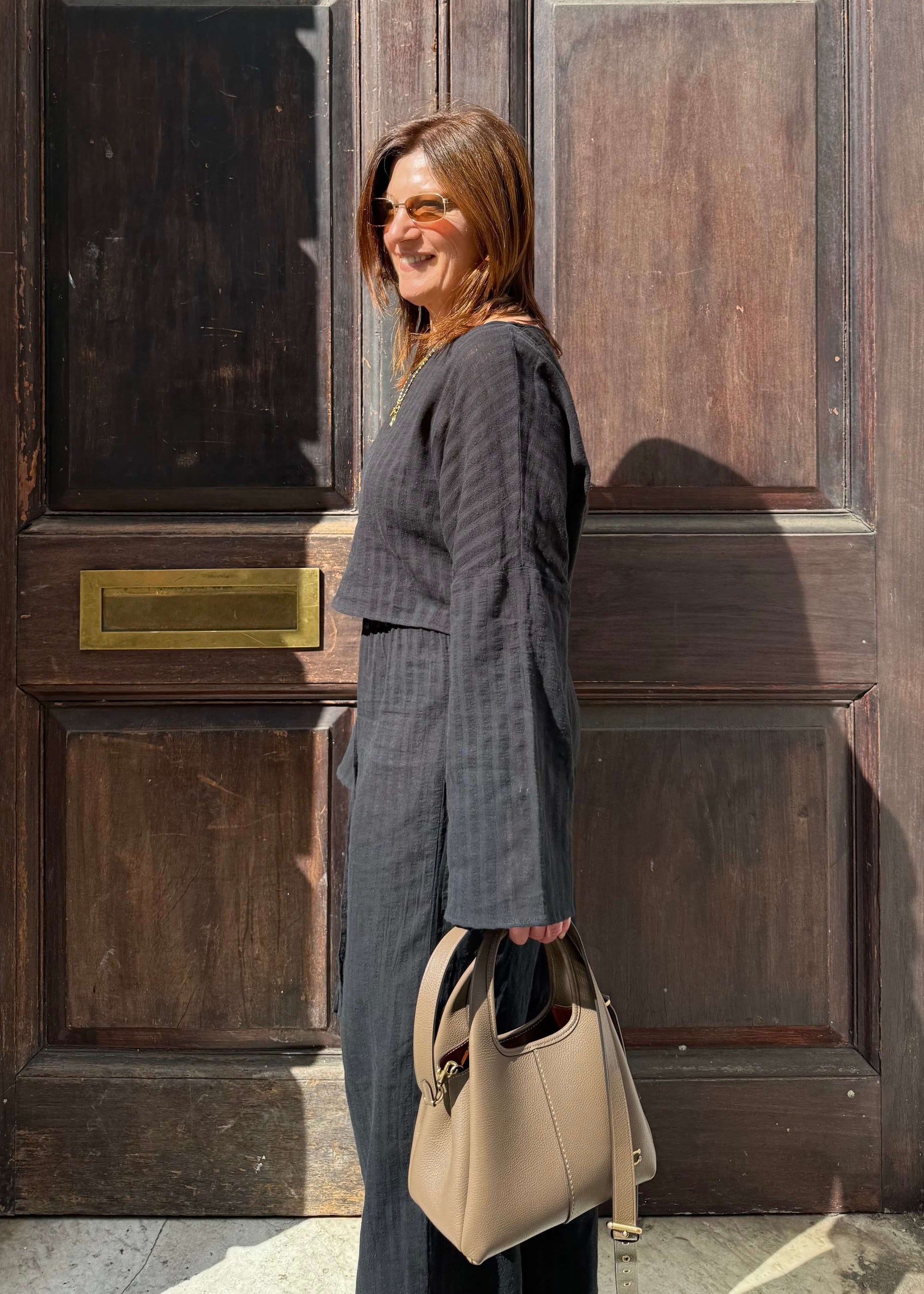 A woman stands in front of a dark wooden door, wearing a black striped two-piece set and holding a handbag.