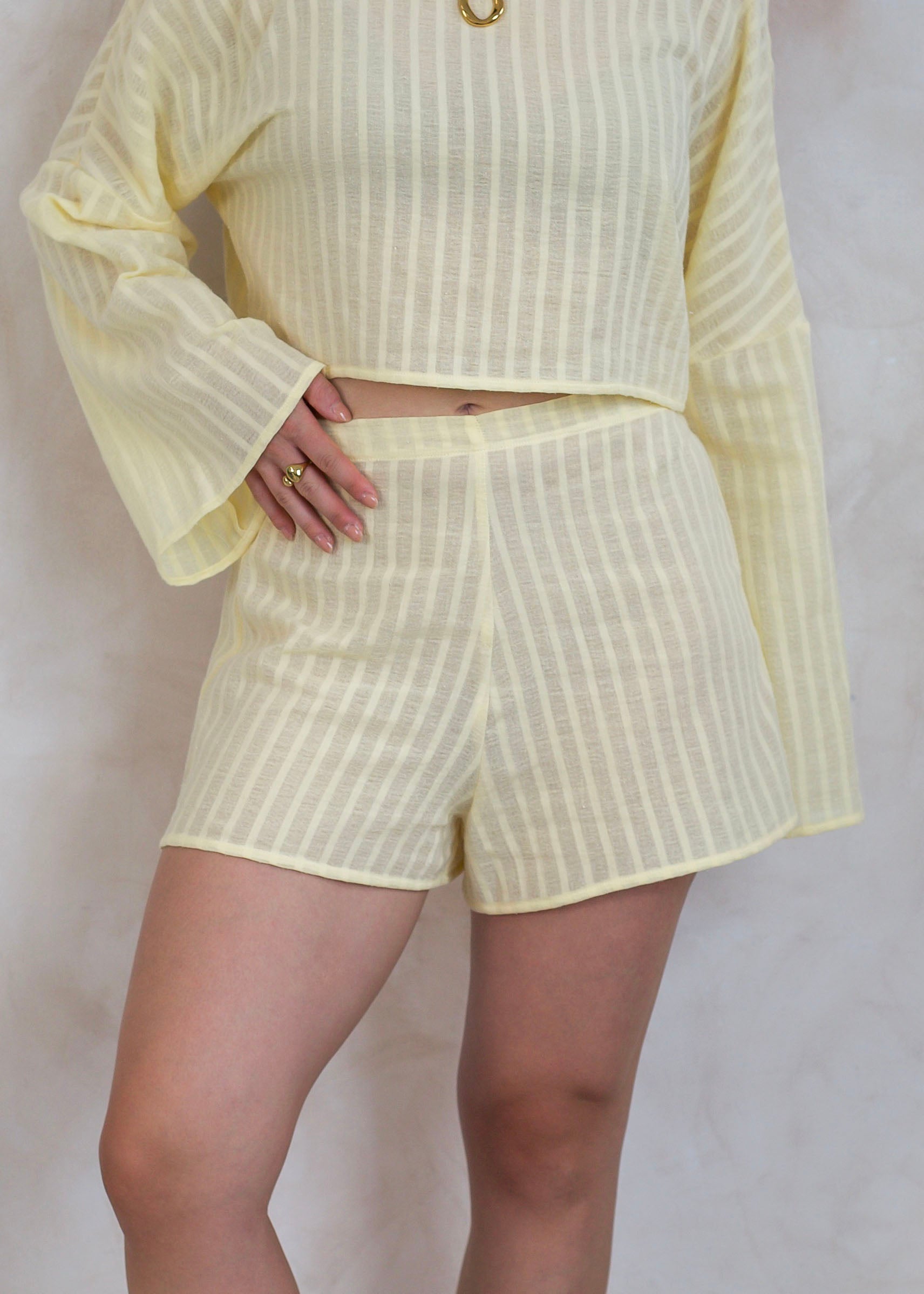 A person wearing a pale yellow two-piece set with vertical stripes, featuring a long-sleeve top and matching shorts, photographed against a light neutral wall.