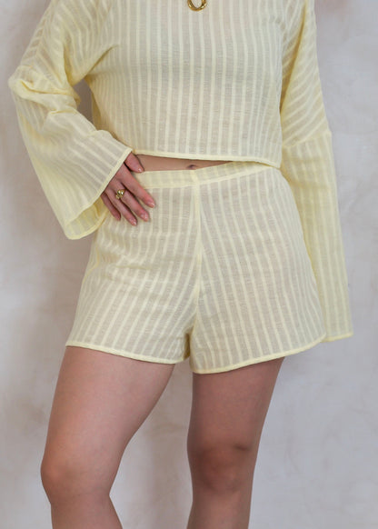 A person wearing a pale yellow two-piece set with vertical stripes, featuring a long-sleeve top and matching shorts, photographed against a light neutral wall.