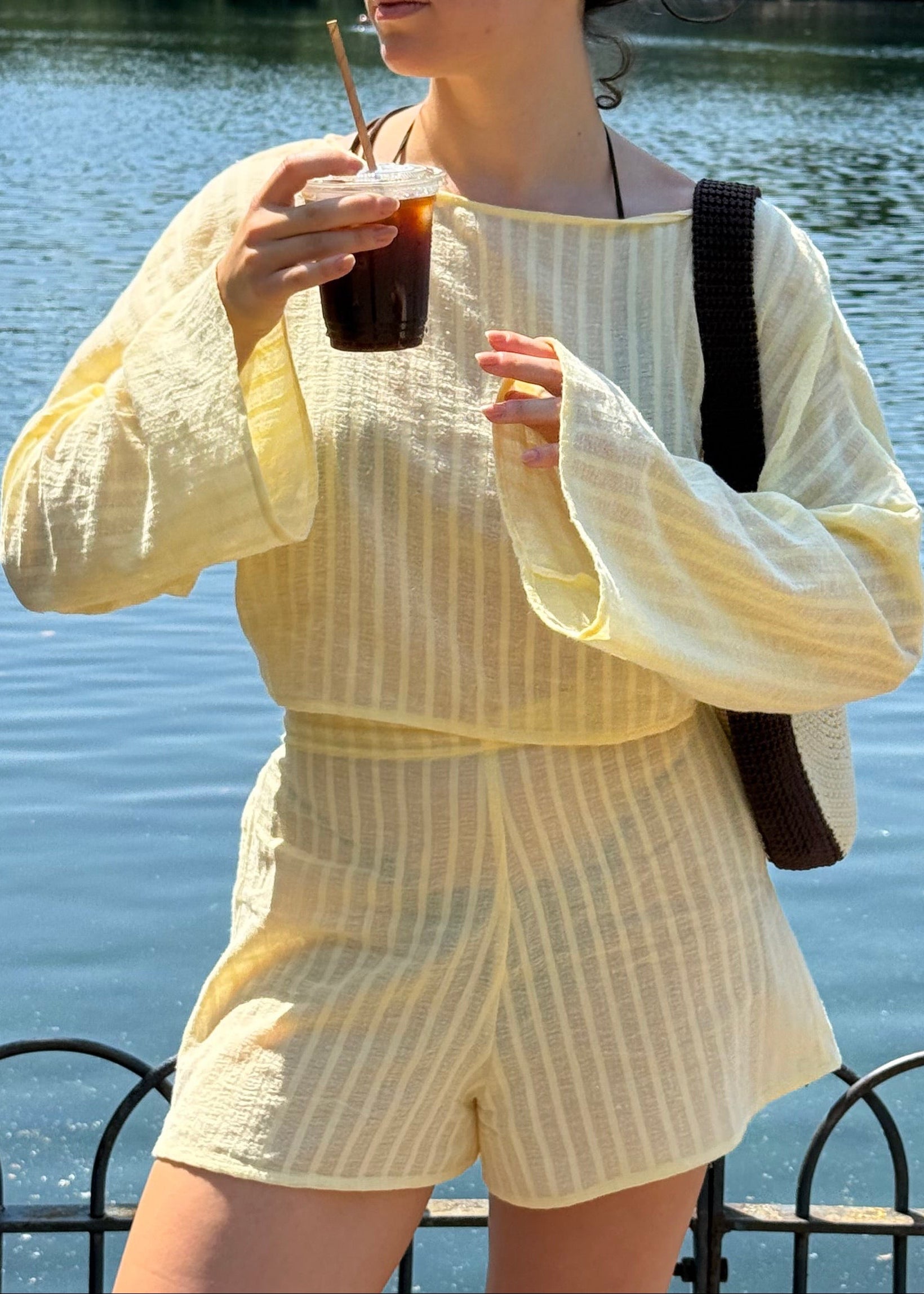 A woman wearing a pale yellow and white vertically striped two-piece set with a boat neckline, long flared sleeves, and matching shorts, standing by a body of water and holding an iced coffee.