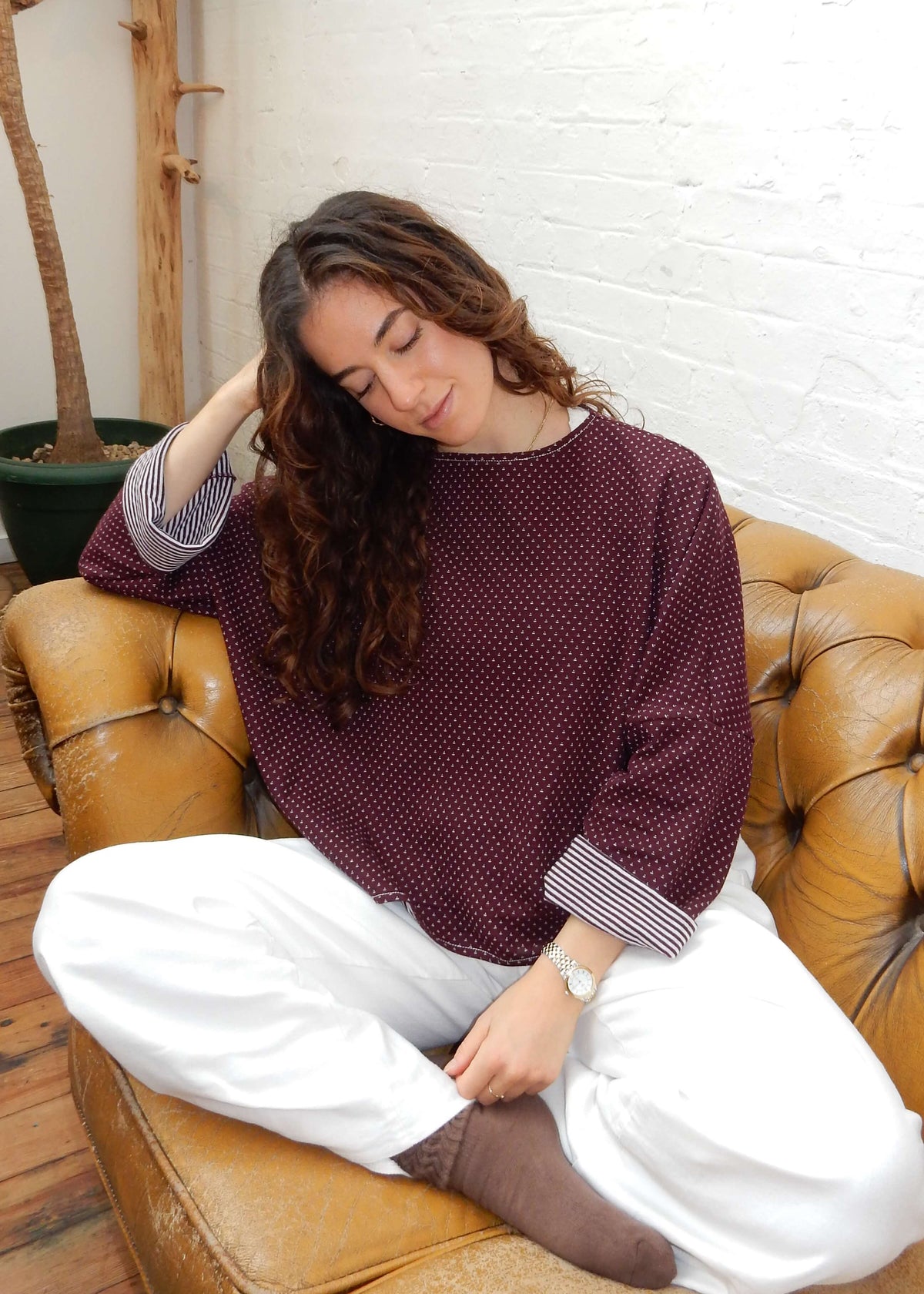 A woman sits cross-legged on a tan leather sofa in a bright room, wearing a burgundy long-sleeve knit with a small white pattern, white jeans, and brown socks.